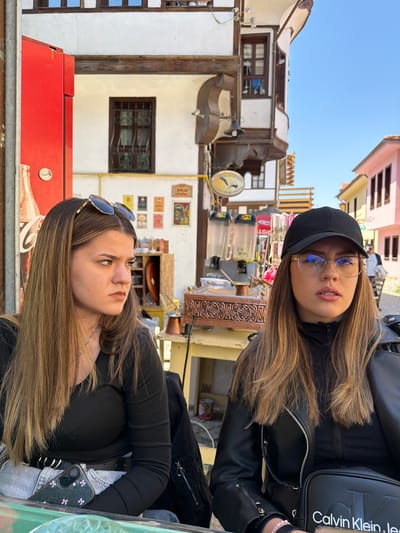 The image captures two young women seated outdoors, likely at a cafe or market stall, in a vibrant, traditional street scene. The original image is rotated 90 degrees clockwise.
In the upper portion of the frame (when upright), a woman with long, light brown hair wears a black baseball cap, round glasses, a black leather jacket over a black ribbed top, and has visible braces. She carries a black shoulder bag prominently displaying "Calvin Klein" in white text. Her gaze is directed slightly upwards and forward, with a neutral expression.
In the lower part of the frame, another woman with long, light brown hair has sunglasses resting on her head. She wears a black long-sleeved top and a silver chain necklace. Her phone, in a dark case adorned with purple floral designs, is partially visible. She looks towards her right, appearing contemplative or observing something in the distance.
The background features multi-storied buildings with traditional architecture, characterized by white walls, wooden elements, and window grilles, suggesting an old town or market area in Eskisehir, Turkey. A bright, clear blue sky is visible above. Various elements hint at commercial activity, including a large red counter or vending machine, decorative wooden carvings, and a circular sign depicting a fish or whale. Several small, framed posters or signs with faded or illegible text are displayed on a wall in the background. The scene suggests a sunny daytime setting, with the women enjoying the ambiance of the locale.