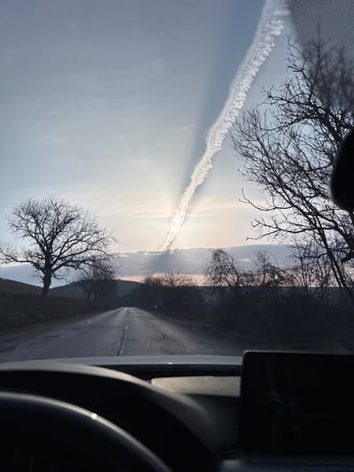 The image captures a rural road scene near Suhache, Bulgaria, viewed from inside a vehicle. The foreground reveals the top of a dark steering wheel on the left and a rectangular infotainment screen on the right side of the dark dashboard, indicating the perspective is through a windshield. An asphalt road, exhibiting cracks and wear, stretches into the distance, marked by a faded white dashed center line and appearing reflective in areas. On the left side of the road, a large, bare deciduous tree stands on a slight elevation, with undulating hills visible in the background. The right side of the road is flanked by a dense line of leafless trees and shrubs, creating a dark silhouette against the brighter sky. Distant hills are faintly discernible on the horizon. The sky is partly cloudy, transitioning from light blue to a pale grey. A bright sun, partially obscured by clouds, is positioned in the middle-left of the upper frame, casting visible crepuscular rays. A prominent, winding white contrail diagonally traverses the sky from the upper right, passing directly through the sun, where it exhibits subtle iridescence. No individuals or vehicles other than the viewpoint vehicle are present.