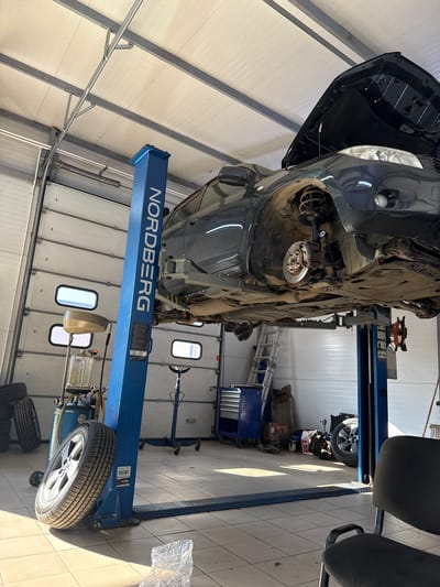 A dark-colored vehicle is elevated on a blue two-post car lift in a garage. The vehicle's hood is open, and the front passenger-side wheel is missing, revealing the suspension and brake components. The garage features white roll-up doors in the background, and various tools and equipment are visible, including stacked tires, an oil drainer, and a toolbox. A ladder leans against a wall, and a chair is situated in the foreground. The scene appears to be a car repair shop.