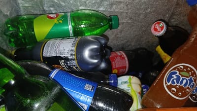 The image displays a collection of plastic and glass beverage bottles accumulated within a grey, possibly insulated, container with an orange border at the bottom right. Condensation is visible on several bottles. The scene is located in Igbede, Nigeria.
Forensic examination reveals the following:
* A green plastic 7UP bottle with its original green screw cap, positioned horizontally at the top left.
* A dark grey plastic bottle, partially compressed, featuring a white label with black text and a checkered pattern, with the letters "ESS" identifiable.
* A dark brown glass bottle with a red and white crown cap, embossed with "Coca-Cola" and an emblem resembling the Nigerian flag, situated in the upper right quadrant.
* A brown plastic Fanta bottle, prominently labeled in blue and white, with "THE COCA-COLA COMPANY" text visible along its side, positioned at the bottom right.
* Multiple dark plastic bottles, one showing a partial blue label containing a QR code.
* Several green glass bottles, some appearing empty, with water droplets on their surface, located on the left side of the frame.
* A small white and green plastic bottle or container marked with "500ml" is partially obscured among other bottles.
* A small, partially visible yellow object is behind the dark brown Coca-Cola bottle.
No individuals are present. The bottles are haphazardly arranged, suggesting a collection of recently consumed or discarded beverages.