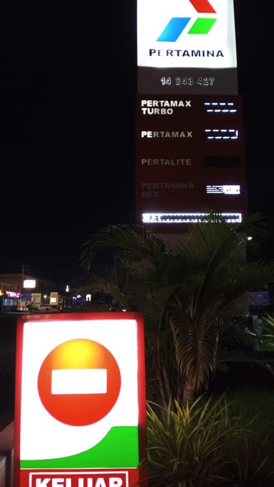 The image shows a night scene at a Pertamina gas station in Cotbadatunong, Indonesia. The main subject is a tall digital price board displaying various fuel types and prices. The Pertamina logo is visible at the top. Below the logo, the number "14 243 427" is displayed. The fuel types listed are PERTAMAX TURBO, PERTAMAX, PERTALITE, and PERTAMINA DEX, each with corresponding prices or symbols indicating pricing. The lower portion of the price board displays "JET." In the foreground, there is a prominent red and white sign with a red circle and a white horizontal bar, symbolizing a "no entry" or "stop" sign, with the word "KELUAR" (meaning "exit" in Indonesian) written below it. Behind the sign, a palm tree and other greenery are visible. In the background, other buildings and lights of the city can be seen. The overall scene is illuminated by artificial lights.