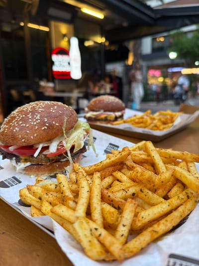 In this close-up, medium-angle shot, a hamburger and a large serving of French fries are presented on a wooden table, likely at an outdoor dining establishment in Izmir, Turkey. The hamburger, in the foreground and slightly to the left, features a sesame seed bun, lettuce, tomato slices, and a beef patty. The fries, seasoned with herbs and spices, are piled high in the lower right quadrant of the image. In the background, slightly out of focus, another hamburger can be seen, along with patrons and the blurred facade of a restaurant with illuminated signage, one of which displays a red burger logo with white text and a white bottle, suggestive of a fast-food or casual dining establishment. The overall setting appears to be a bustling urban environment, characteristic of a street-level eatery in a Turkish city like Izmir.
