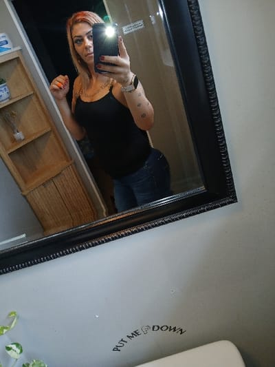 An adult female is depicted taking a mirror selfie in a bathroom. She has long, light-colored hair with visible orange or red highlights. She wears a black tank top, dark blue jeans, and a silver-colored watch on her left wrist. Tattoos are present on both arms, with geometric symbols visible on her right forearm. Her fingernails are painted dark red. She holds a black smartphone with her right hand, positioned to capture her reflection, with the phone's flash activated and illuminating her face. Her left hand is raised slightly, fingers curled.

The mirror has a thick, dark, ornate frame. Reflected in the mirror behind the individual is a wooden shelving unit containing various objects, including a white ceramic pot with blue patterns holding a small plant, and a clear vase with decorative sticks. A faint white label or sticker is visible on a surface reflected behind her left shoulder.

Outside the mirror's reflection, the visible environment consists of a light gray-beige painted wall. A black text decal or sticker is affixed to the wall below the mirror, reading "PUT ME 🚽 DOWN," with a toilet seat icon. A white porcelain object, consistent with the top of a toilet, is partially visible below this decal. A variegated green and white plant is partially visible in the lower left foreground. The specific location within Redondo Beach, United States of America, cannot be determined from the provided image.