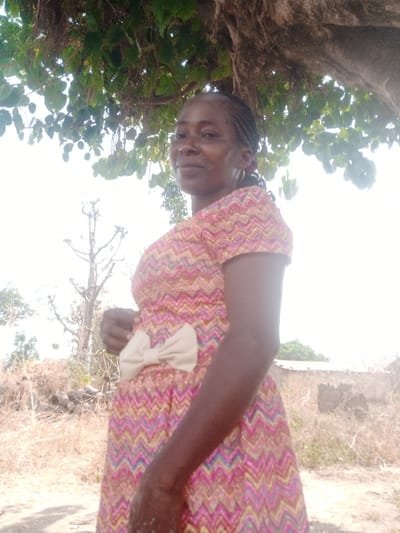 An adult female, with dark skin and braided hair, stands outdoors in Gudige Sabo, Nigeria. She wears a short-sleeved dress patterned with multicolored zigzags (pink, yellow, blue, green) and a prominent beige bow at the waist. She faces the camera with a slight smile, her right hand resting near the bow. Overhead, green tree foliage provides partial shade. The background features dry earth with sparse, dried vegetation, a tall leafless tree structure to the left, and a low, unfinished concrete or mud wall structure to the right. The sky is bright and clear, indicating strong daylight conditions.
