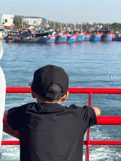 In the city of An Hải, Vietnam, a young boy wearing a black t-shirt and a black baseball cap is seen from behind, leaning on a bright red railing. He is facing a body of water where numerous colorful fishing boats are docked in the background. The water is a vibrant blue, reflecting the clear sky above. Buildings and lush greenery can be seen on the shoreline behind the boats. The overall scene suggests a sunny day at a bustling harbor.
