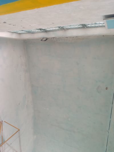 The image displays an interior view of what appears to be an unfinished or disused room. The walls are painted a light, possibly pale blue or green, with visible water stains and some unevenness in the paint application. In the upper portion of the image, a yellow band runs horizontally across the top edge, above a white ceiling that shows some wear and tear, including dark spots and a small section of exposed metal grating. This grating appears to be part of a ventilation or light fixture.
In the lower left corner, a portion of a structure with a wooden frame and what seems to be glass or transparent panels is visible. This could be a shelf, a display case, or part of an unfinished piece of furniture. The overall impression is of an interior space that is either under construction, undergoing renovation, or has been left neglected. There are no people visible in the image, nor any clear indication of recent activity. The lighting suggests it is daytime, possibly with natural light entering from the grating in the ceiling. The scene is devoid of any clear landmarks that would specifically place it within Boroldoy, Kyrgyzstan, beyond the provided context.