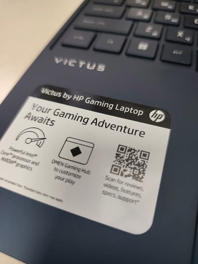 The scene is situated in the city of Kangli, Uzbekistan. The image provides a close-up, angled view of a dark blue HP Victus gaming laptop. The silver "VICTUS" logo is visible on the laptop's palm rest. A portion of the dark-colored keyboard with square keys is blurred in the upper right background. A prominent white, rectangular sticker with rounded corners is affixed to the laptop. The sticker's upper section reads "Victus by HP Gaming Laptop" alongside the HP logo, followed by the phrase "Your Gaming Adventure Awaits". The lower section of the sticker contains three informational graphics: a speedometer-like icon with text "Powerful Intel® Core™ processor and NVIDIA® graphics"; an icon of a diamond within a box, labeled "OMEN Gaming Hub to customize your play"; and a QR code with accompanying text "Scan for reviews, videos, features, specs, support*". The laptop rests on an undifferentiated light-colored surface. No individuals are present, and no actions or interactions are depicted.