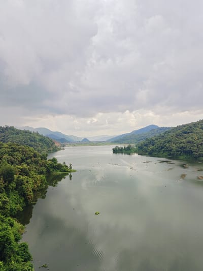This is a wide shot, taken from a high vantage point, looking out over a large, calm lake in Rupakot, Nepal. The sky is overcast with soft, diffused light. The lake dominates the lower half of the frame, its surface reflecting the cloudy sky and the surrounding green hills. The water exhibits gentle ripples and there are scattered small islands or debris visible on its surface.
To the left of the lake, lush green trees densely cover the sloping hillsides, their vibrant foliage creating a rich texture. A small boat can be seen on the lake in the middle ground, heading away from the viewer towards the right. In the distance, rolling hills, also covered in trees, recede into the hazy background, with hints of human settlement and agricultural fields visible between the natural landscape. The overall scene is serene and natural, with the overcast sky contributing to a peaceful, almost muted atmosphere.