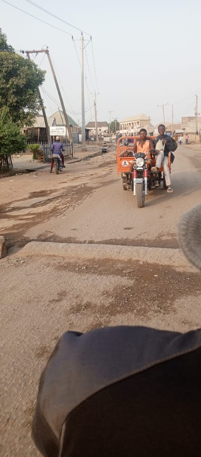 The image captures a daytime street scene in Tunga Mekidi, Nigeria. An orange three-wheeled motorcycle (keke napep) with two young male occupants is prominent in the mid-ground, traveling towards the viewer on a paved road. The male operating the vehicle wears an orange tank top, while his passenger, seated in the cargo area, wears a black jacket and light trousers, and is looking towards the camera with a smile. The road surface is asphalt, showing wear with visible cracks and uneven patches, and features raised concrete barriers or speed bumps.
On the left side of the road, a second individual wearing a blue patterned shirt and a light-colored helmet rides a two-wheeled motorcycle away from the camera, passing a sign for "PARACLET RAPHA HOSPITAL LTD" in front of a building with a green roof and a metal fence. The surrounding environment includes various utility poles with overhead wires, green foliage from trees and shrubs, and multiple single and multi-story buildings lining the street in the background. The sky is clear and light. The bottom foreground shows a blurred dark surface, consistent with the photographer's immediate surroundings.