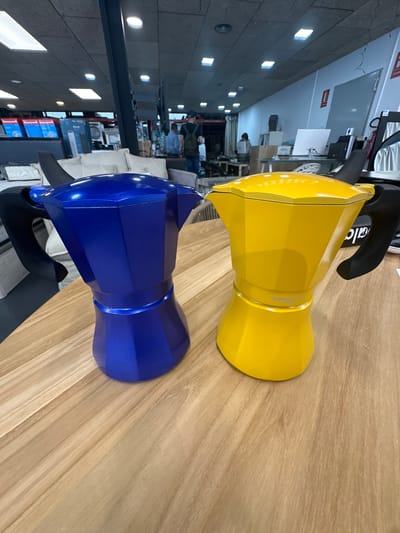 Two vibrant Moka pots, one yellow and one blue, are prominently displayed on a light-colored wooden surface with a distinct grain. The image is oriented vertically, making the Moka pots appear to be lying on their sides. Both coffee makers feature a classic octagonal design with black handles. The yellow Moka pot has "D'ORO" subtly inscribed on its body and part of the "Bialetti" brand name visible on its black handle. The blue Moka pot has a metallic sheen.
The setting appears to be an indoor commercial space, possibly a showroom or store in Andorra la Vella, Andorra. The background is intentionally blurred, directing focus to the Moka pots. Visible in the soft-focus background are various elements indicating a retail or office environment. These include shelves, boxes (some clearly marked "Honeywell"), computer monitors, whiteboards, and diverse lighting fixtures like square and round ceiling lights. A concrete or stone-like wall is visible on the left side of the frame. Blurry figures of people can be seen in the distance, some seemingly working on laptops or browsing, suggesting typical daytime activity within the space. A red sign with "EXTINTOR" (fire extinguisher) and another small sign marked "EI" are also discernible on the back wall. The overall impression is that of products showcased in a modern retail or display area.