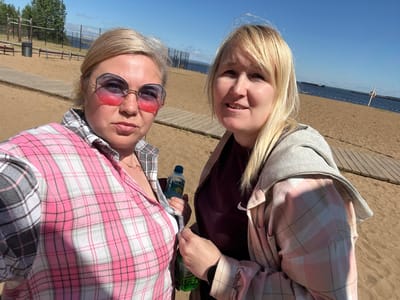 Two women are captured in a selfie on a sandy beach, with a clear blue sky overhead. The woman on the left, wearing large, gradient pink-to-blue sunglasses and a pink, white, and grey plaid shirt, holds a bottle of water. The woman on the right, with blonde hair, wears a burgundy shirt and a light grey hooded jacket over a similar pink plaid shirt, and holds a green bottle. Wooden walkways are visible on the sand behind them, leading towards the water. In the background, a fence, some trees, and distant structures are present, suggesting a recreational area. The overall scene depicts a casual outdoor outing. The location is identified as the city of Galevo, Russia.
