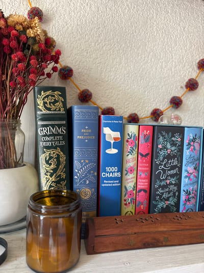 A stack of books sits beside a wooden box and an amber glass jar. Book titles visible include "Grimm's Complete Fairy Tales," "Pride and Prejudice," "1000 Chairs," "Heidi," "A Little Princess," and "Little Women." A garland of pom-poms hangs nearby, and a vase of dried flowers sits at the base of the books. The arrangement is casual, with a focus on literary classics and decorative elements.