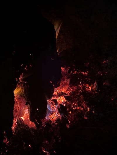 The image shows a close-up view of glowing embers and burning wood in what appears to be a campfire. The scene is predominantly dark, with the bright oranges, reds, and hints of blue from the fire illuminating the charred wood and ash. The texture of the wood is rough and gnarled, with cracks and crevices where the heat is most intense. Scattered embers, appearing as small, bright sparks, are visible throughout the scene, especially in the lower right portion. The background is almost entirely black, suggesting a nighttime setting. There are no discernible people or identifiable objects beyond the fire itself. The overall impression is one of intense heat and decay. The location is specified as Bliznatsi, Bulgaria, but this contextual information cannot be directly verified from the visual content of the image alone.