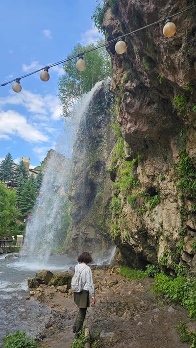 In the city of Krasny Kurgan, Russia, a young person with dark, curly hair and a light gray backpack is standing on a muddy path, facing a waterfall. They are wearing a light gray sweatshirt and an olive-green skirt or dress over olive-green leggings or tights. The waterfall cascades down a large, rocky cliff face that is covered in green vegetation. In the background, to the left of the waterfall, are evergreen trees and a building with a tower, resembling a castle. Above the scene, a string of round, warm-toned lights is strung across the frame against a bright blue sky with scattered white clouds. The ground in front of the waterfall is a mix of rocks and mud, with a stream flowing to the left.