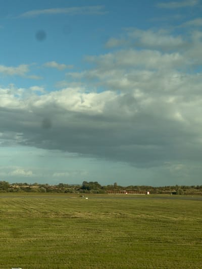 The image depicts an airfield landscape under a partly cloudy sky, located in Mitry-Mory, France. The foreground consists of a wide expanse of mown green grass, exhibiting distinct parallel striations from maintenance. Beyond the grass, a dark grey paved surface, likely a runway or taxiway, traverses the mid-ground horizontally. Several small white rectangular markers are positioned on or adjacent to this paved area. In the background, a dense line of green foliage, comprising trees and bushes, stretches across the horizon. Above and partially obscured by this foliage, a series of red-framed structures with white bases, possibly approach lights or navigational aids, are visible along the paved surface. The sky occupies the upper two-thirds of the frame, transitioning from clear blue in the upper left to a mix of white and dark grey cumulus and stratus clouds dominating the central and lower sky. Two distinct circular dark smudges, consistent with sensor dust, are present in the upper and middle sections of the sky. No individuals or active subjects are discernible within the scene.