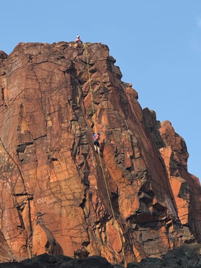 In Valok, Russia, two individuals are engaged in rock climbing activities on a steep, reddish-brown rock face. The first climber, wearing a blue jacket and a helmet, is positioned mid-climb, attached to a yellow rope. Above and to the left, a second climber, also wearing a helmet and a pink jacket, is seated at the summit of the rock face. A red rope is visible extending down the right side of the rock face. The rock face is textured with cracks and appears to be a natural formation, with a clear blue sky serving as the backdrop. The ground below the rock face is covered in dark, rough terrain.