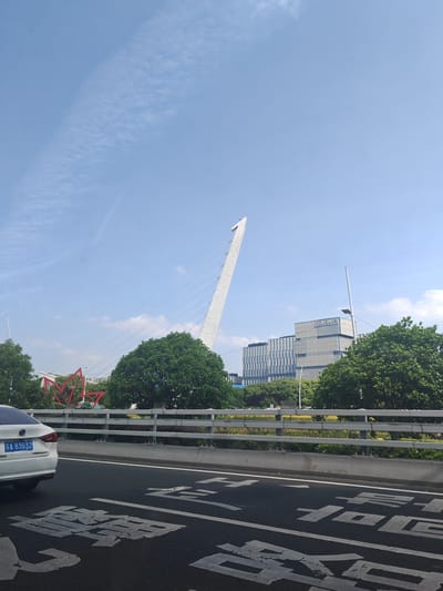 A low-angle shot taken from a vehicle captures a daytime scene in Qinhuai District, China. The vehicle, with license plate 苏A 83G32 visible, is traveling on a road with painted white Chinese characters. A grey metal guardrail separates the road from a green area with trees and landscaping. A white cable-stayed bridge is the central focus, with the building of the Jiangsu Academy of Traditional Chinese Medicine and some red lotus-shaped decorative elements in the background. The sky is mostly blue with some faint clouds.