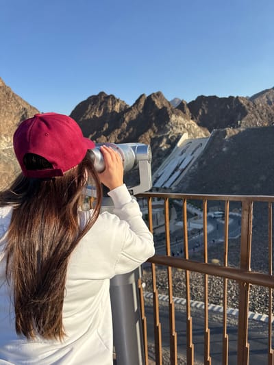 A person is observed from behind on an elevated viewing platform in Hatta, United Arab Emirates, specifically overlooking the Hatta Dam area. The individual, a female with long dark brown hair, wears a burgundy baseball cap and a white long-sleeved top. She is actively using a silver, stand-mounted public binocular telescope, holding its eyepiece with her right hand and looking out towards the landscape.
The scene unfolds under a clear blue sky, revealing rugged, brown and grey mountainous terrain in the distance. To the right of the frame, a significant concrete dam structure is visible, with large white letters spelling "HATTA" prominently displayed on its sloped face. Below the viewing platform, a brown metal railing spans the foreground. Beyond this railing, a winding road is discernible, with small figures (people) and a white vehicle visible, indicating human activity along the route. The environment is bright, suggesting daylight conditions.