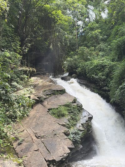 A lush green jungle scene surrounds a cascading waterfall at Ban Huai Takhrai, Thailand. The waterfall flows over large, weathered rocks, with water splashing and creating foam at the bottom. Dense vegetation, including trees, vines, and various plants, covers the surrounding cliffs and hillsides, creating a vibrant, natural environment. Sunlight filters through the canopy, illuminating parts of the scene with a soft glow. The image captures the raw beauty of the natural landscape.
