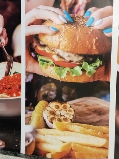 The image features a close-up of a burger held by hands with turquoise and silver nail polish. The burger contains a sesame seed bun, lettuce, tomato, cheese, and other fillings. Below the burger, on a wooden board, are french fries, a pickle, and roasted garlic. To the left, a separate plate shows spaghetti with tomato sauce. The focus is on the burger and its accompanying sides.