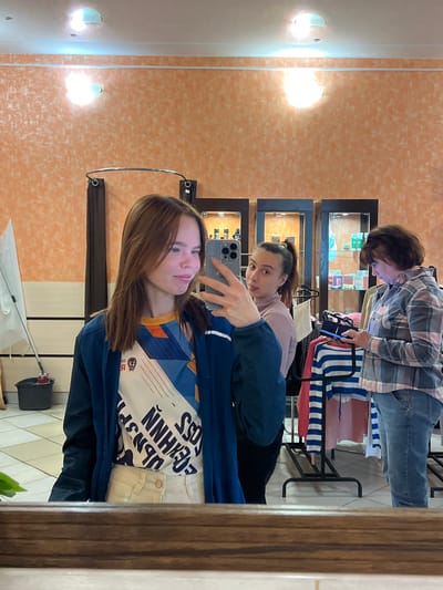 The image is a selfie taken in a commercial establishment, likely a store or boutique, in Bol'shaya Sosnova, Russia. The main subject is a young woman holding a smartphone (likely an iPhone based on the camera configuration) to capture a reflection in a mirror. She is wearing a blue jacket over a patterned top and light-colored pants. Behind her, another woman with her hair pulled back into a ponytail looks towards the camera, seemingly observing the selfie being taken. Further in the background, an older woman wearing a plaid shirt looks down, potentially examining merchandise in a display case. The store appears to sell small goods or accessories, as evidenced by the display cases. A chair with striped upholstery is also visible. The store walls are textured and painted a pale orange color. There are illuminated wall sconces on the wall near the mirror.