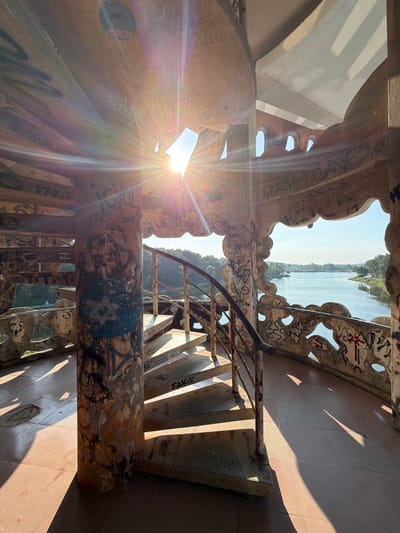 This image depicts the interior of the unfinished Hồ Thủy Tiên Water Park in Thủy Xuân Ward, Vietnam, specifically the inside of a decaying architectural structure, possibly a lookout tower or part of a ride. A concrete spiral staircase with rusted metal railings ascends through the interior. Graffiti covers the majority of the concrete surfaces. Natural light streams in from above and from an opening looking out onto a body of water, possibly a lake or river. The light creates strong lens flares and casts harsh shadows. The exterior landscape is visible in the background, showing green vegetation and a water expanse. The floor is tiled.