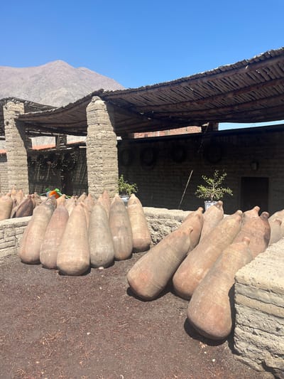 The image shows the exterior of a structure with a weathered wooden roof and a brick and cement facade. A mountain range is visible in the background under a clear blue sky. Rows of large, earthenware jars are stacked along the side of the building. The ground around the structure is covered in dirt and debris. The location is the city of Lunahuaná, Peru.