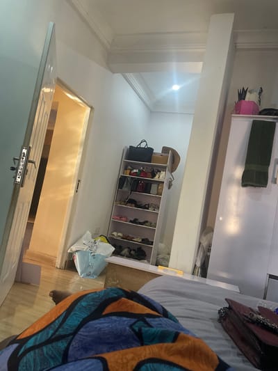 A photograph depicts a room interior, likely a bedroom or dressing area. The room is predominantly white, with a white wardrobe and a shoe rack. There are multiple pairs of shoes displayed. A door is visible in the image, and a light is shining through the doorway. Part of a person wearing a blue and orange patterned garment is in the foreground. The presence of indoor footwear and personal items suggests a private residential space. The image's location is unknown, and there is no way to verify that it is in the city of Dushepe, Nigeria.