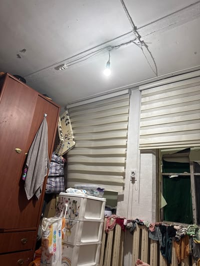 The image shows an indoor scene, likely a cluttered room. The subjects are primarily objects and the setting itself. The scene is dimly lit by a single bare light bulb hanging from the ceiling. Wires are visible, running along the white ceiling and walls. There are vertical blinds covering windows or sections of the wall. In the foreground, a dark wooden dresser or cabinet is visible, with a folded gray towel and a patterned cloth draped over it. Behind the dresser, there are stacked items, including what appear to be fabric storage cubes with animal prints and a package of diapers. Clothing items, possibly socks, are strung up along a window frame or ventilation duct on the right side of the image. The overall impression is one of disarray and utilitarian storage. There is no visible indication of the time of day or weather. The location context provided is Shamalgan, Kazakhstan, but no specific visual cues within the image confirm this. There is text visible on the diaper packaging, including "Happy Star" and size indicators like "S 50". Other text on the packaging is too small or blurry to be legible.