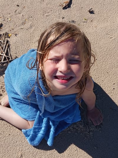 The image displays a young child, appearing to be female, sitting or kneeling on a sandy surface in the city of Podborov'e, Russia. The child's light brown hair is wet, with individual strands visible. She is wrapped in a textured, light blue terry cloth towel that covers her torso and legs. Her eyes are squinted, suggesting exposure to bright sunlight, and she exhibits an open-mouthed smile, revealing both upper and lower teeth. Her cheeks appear rosy.

The environment consists of light tan, fine-grained sand, sparsely scattered with small pieces of natural debris such as dried twigs and small, dark organic matter. Strong, defined shadows are present, with a significant dark shadow extending across the lower right portion of the frame, indicating intense, direct sunlight from an overhead or elevated source. The child's visible right hand is positioned in the lower right, partially gripping the sand. Her left leg is bent, with the knee visible beneath the towel.