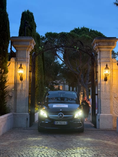 A black Mercedes-Benz minivan with headlights illuminated is parked at the entrance of a stone gate, illuminated by two ornate lanterns. The minivan has a license plate displaying "GM-782-DL". A woman in a black dress is standing next to the open driver's side door, holding a smartphone. The gate is adorned with decorative ironwork and flanked by large stone pillars and tall cypress trees. The ground is paved with cobblestones. The scene appears to be in the city of Vallauris, France, at dusk.