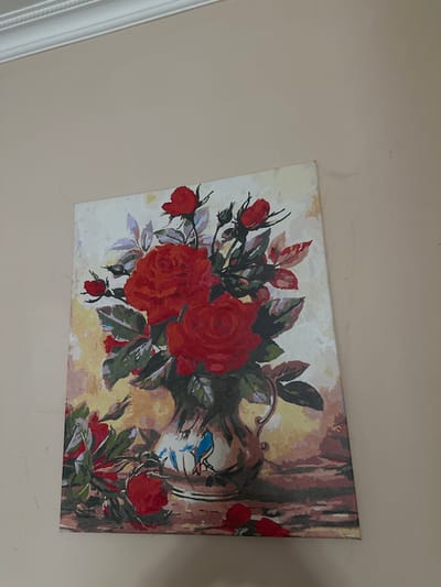 The image shows a painting of a vase of red roses. The painting is hanging on a light-colored wall, and there is decorative molding at the top of the wall. The painting appears to be a still life, with the roses arranged in a vase on a table. The background of the painting is a soft, muted yellow and brown. There are several red roses, some in full bloom and others as buds. There are also green leaves and stems. A blue bird is depicted on the vase. The painting has a textured appearance, suggesting it might be a print or a painting by numbers. The scene is static and does not depict any action or interaction. The location is not specified in the image, and therefore cannot be determined.