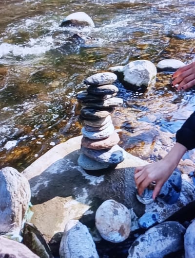 The image depicts a close-up scene at a river or stream bank in Aktash, Uzbekistan. In the central foreground, a cairn composed of approximately 11-12 smooth, water-worn stones of varying sizes and muted colors (greys, browns, and blues) is stacked vertically on a larger, flat rock. A human hand, with dark nail polish, is visible in the lower right portion of the frame, reaching towards smaller, wet, rounded rocks on the bank. A second human hand, also with dark nail polish, is partially visible further back in the water, near submerged rocks. The background consists of flowing water with visible ripples and areas of foam, indicating current. Numerous rounded, submerged, and partially submerged rocks are visible through the clear, brownish-green water. The scene is illuminated by natural light.