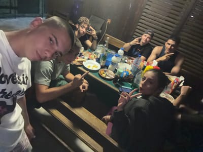 In Vladislavtsi, Bulgaria, a group of young adults is gathered around a table, likely for a meal or social event. The scene is outdoors, possibly a patio or covered area, with wooden slatted walls and a roof visible. A table is laden with food, plates, bottles of drinks, and what appears to be a water pitcher. The individuals, all appearing to be in their late teens or early twenties, are engaged in various activities. One person in the foreground is taking a selfie. Others are interacting with each other, some making gestures with their hands. The lighting is dim, suggesting it is evening or nighttime. The overall atmosphere seems casual and informal.