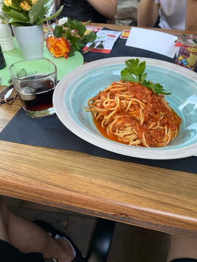 The image shows a plate of spaghetti bolognese on a table in what appears to be an outdoor seating area of a restaurant. The spaghetti is topped with a hearty meat sauce and garnished with a sprig of parsley. To the left of the plate is a glass of dark liquid, possibly cola or beer, and a small vase with a yellow and orange rose. In the background, out of focus, are people and a jar of colorful candies. The table is made of wood, and there is a black placemat under the plate and glass. The overall impression is a casual dining setting. The provided information states that the location is in Wölfersheim, Germany, which is a municipality in the Wetterau district of Hesse. This suggests the restaurant is a local establishment in that area.