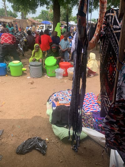 This media file captures a vibrant outdoor market scene in Nigeria. The foreground is dominated by colorful fabrics, some appearing to be clothing or textiles, hanging from an overhead structure, creating a canopy. Below, a diverse group of people are seen engaged in various activities. Some are seated, possibly vendors or shoppers, while others appear to be standing or moving through the market. Several motorcycles are parked, suggesting transportation is a key element of the setting.
Various objects are scattered throughout the scene, including bright green plastic buckets, grey containers, and what appear to be water jugs or storage bins. A black plastic bag lies on the ground in the lower right corner, hinting at the presence of waste or discarded items. In the background, simple structures, likely market stalls or homes, are visible, along with trees and vegetation, indicating a rural or semi-urban environment. A white truck with an umbrella on top adds to the bustling atmosphere.
The lighting suggests it might be daytime, possibly late morning or early afternoon, with the sun casting shadows. The overall mood appears to be one of daily life and commerce, with people actively participating in the market.
Visible text in the image includes brand names such as "Calvin Klein" and "BJÖRN BORG" repeated on fabric straps or ribbons, possibly part of the merchandise being sold. The scene depicts a typical day at a Nigerian market, alive with activity, trade, and community interaction.