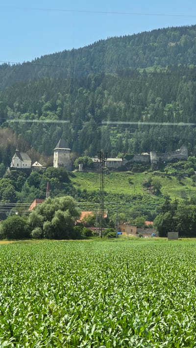 A wide-angle, eye-level shot captures a verdant cornfield in the foreground, stretching towards a backdrop of a lush green hillside. Perched atop the hill are the remnants of a historic castle and a small white church with a red-roofed steeple. The castle, with its weathered stone walls and towers, appears to be partially in ruins, hinting at centuries of history. The church, in contrast, stands pristine and white, its steeple reaching towards the clear blue sky. Scattered amongst the greenery are several houses, some with red roofs, and a tall electrical tower, suggesting the presence of modern infrastructure within this picturesque setting. The distant hills are densely forested, creating a dramatic contrast with the open fields in the foreground. The image appears to be taken from a moving vehicle, possibly a train, as indicated by the faint horizontal lines that run across the frame, suggesting the presence of windowpanes. The overall impression is one of a tranquil rural landscape, where history and nature coexist harmoniously. The specific location is the historic town of Friesach, Austria.