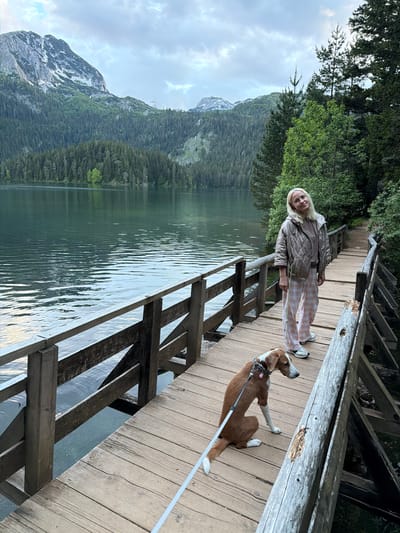 This image depicts a scene in Pitomine, Montenegro, featuring a wooden boardwalk running along the shore of a serene mountain lake. A blonde-haired woman stands on the boardwalk, facing the viewer with a slight smile. She wears a quilted beige jacket, a light brown t-shirt, and pink and white plaid trousers with light-colored athletic shoes. To her left, a brown and white dog with short fur sits on the wooden planks, looking towards the right side of the frame. The dog is attached to a white leash held by the woman and wears a metal chain collar.
The wooden boardwalk, constructed from aged timber, includes a simple railing system along the lake's edge. The lake exhibits a dark greenish hue, with gentle ripples disturbing its surface, reflecting the cloudy sky. Across the lake, dense evergreen forests cover the lower slopes, transitioning into rugged, grey mountain peaks, some of which bear patches of snow. The sky is overcast with grey and white clouds. To the right of the woman, a natural dirt path extends into a thick forested area, indicating a surrounding wilderness. The overall setting is a natural landscape with mountainous and aquatic features.