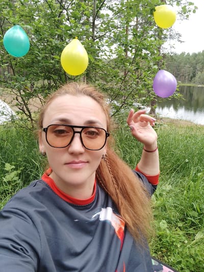 A woman with long, reddish-brown hair and sunglasses is taking a selfie outdoors. She is wearing a dark grey shirt with a red collar. She has a bracelet on her wrist. In the background, there are trees, green grass, and a lake. Four balloons are hanging from a tree: one teal, one yellow, one purple, and another yellow balloon. The scene appears to be a park or natural area. The lighting suggests it is daytime, and the cloudy sky indicates potentially overcast weather. The woman's expression is neutral.