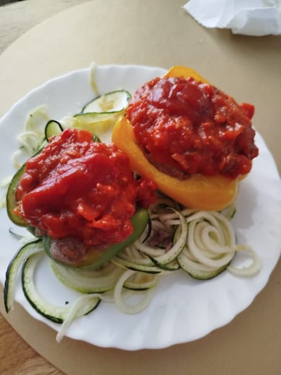 Two stuffed bell peppers, one green and one yellow, are presented on a white, scalloped-edge plate. The peppers are filled with a meat mixture and topped with a chunky tomato sauce. Surrounding the peppers are spirals of zucchini, some with green skins and some with white flesh. The plate sits on a light brown, textured surface, possibly a wooden table. A partially visible white paper napkin is in the upper right corner of the image. The scene appears to be indoors, likely a home kitchen or dining area, as there are no distinctive external features indicating a specific location in Offenbach am Main, Germany.