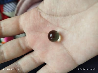 A single, dark, gelatinous capsule, approximately 0.5 cm in diameter, rests on the palm of a person's hand. The capsule has a glossy surface and appears to be translucent, with a darker core. The hand is fair-skinned with visible lines and creases. The background is blurred, showing hints of fabric in shades of pink, blue, and red. The scene is illuminated by artificial light. The date stamp "19.06.2026 10:57" and "REDMI NOTE 13" are visible in the lower corners. The location is not determinable from the image.