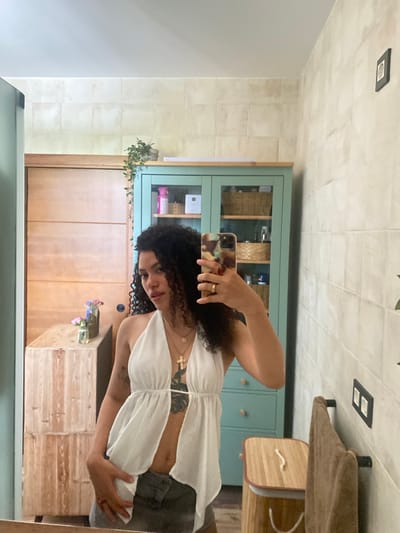A woman with curly dark hair, wearing a white halter top and grey shorts, takes a mirror selfie in a bathroom. She is holding a mobile phone in her right hand and has a ring on her left hand. She is wearing a necklace with a cross pendant. In the background, there is a wooden cabinet, a blue cabinet with shelves, and a towel hanging on a rack. The walls are tiled. The location is the city of Zeneta, Spain.