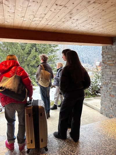 A group of five individuals stands in a sheltered outdoor alcove with a wooden plank ceiling and a speckled concrete floor, looking out at a snowy, mountainous landscape in what is identified as La Clusaz, France. To the right, a wall is clad with stacked grey stone. Snow covers the ground beyond the alcove, and numerous evergreen trees are visible, with distant buildings nestled on snow-covered slopes.

From left to right:
1.  A person in a bright red puffy winter jacket, blue jeans, and pink footwear, seen from behind, carrying a dark grey shoulder bag with a gold chain strap. A light gold wheeled suitcase stands upright next to their left leg.
2.  An individual, viewed from behind, wearing a dark vest over a long-sleeved top and blue jeans, carrying a beige backpack with "A.P.C." visible.
3.  A person in a dark puffy winter jacket, light grey sweatpants, and dark boots, with blonde hair, partially obscured.
4.  A younger individual with dark hair, seen in profile, wearing a dark top, positioned behind the person in the black jacket.
5.  A person with long dark hair, seen from behind, wearing a black puffy winter jacket, black wide-leg pants, and dark footwear.

The individuals appear to be observing the snowy alpine environment. The sky is overcast.