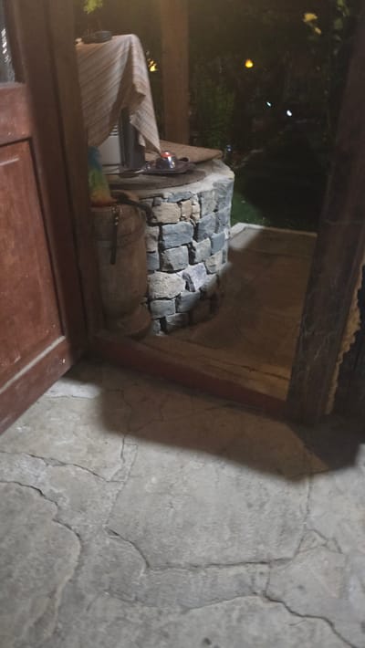 The image depicts an outdoor scene at night, possibly in Kaleytsa, Bulgaria. In the foreground, a cracked concrete surface is visible. To the left, a wooden door with vertical planks is partially open. In the center, a stone structure, possibly a well or decorative feature, is prominent. This structure is made of rough-hewn stones and is topped with a blanket draped over an object. A small tray with what appears to be food or a candle is also on top of the stone structure. A wooden post and foliage are visible in the background, illuminated by ambient lighting, with a dark, shadowy garden area beyond. The scene conveys a rustic, possibly natural setting at dusk or night.