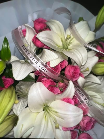A close-up, slightly blurry image shows a bouquet of white lilies and pink roses. A ribbon with the words "JUST FOR YOU" printed in black lettering is draped across the flowers. The bouquet is wrapped in white paper. There are no people visible in the image. The setting appears to be indoors, possibly a gift shop or a home, and there is no indication of a specific location in Kadamjay, Kyrgyzstan.