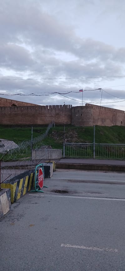 The image shows a section of the Ivangorod Fortress in Ivangorod, Russia, located along a road. A stone wall with battlements is visible behind a metal fence topped with barbed wire. A stop sign is positioned on a barrier adjacent to the road. The sky is overcast.