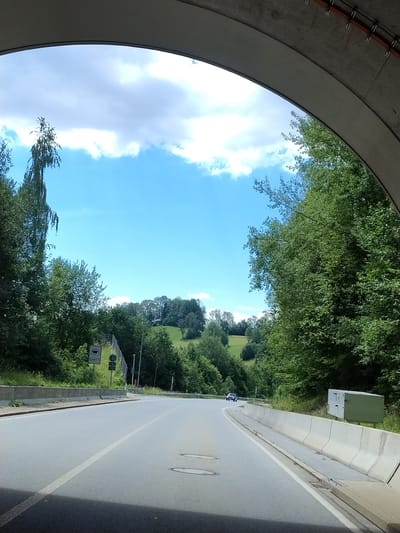 The image shows a view from inside a tunnel exit, looking out onto a road bordered by trees and a grassy hillside. The sky is blue with some clouds. A road curves to the right, with a white line marking the lanes. There is a speed limit sign with a pictogram of a car. On the right side of the road, there is a concrete barrier and a metal box. In the distance, a dark blue car is driving away from the camera. The scene appears to be a rural area, possibly near Röhrnbach, Germany, given the context.