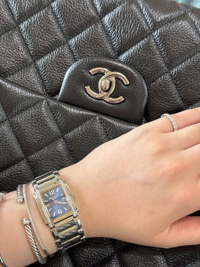 A close-up shot focuses on a person's hand and wrist adorned with jewelry, resting against a black quilted handbag with a prominent interlocking "C" logo. A diamond ring is visible on the ring finger. A silver-toned wristwatch with a rectangular blue face is positioned on the wrist, along with two silver-toned bracelets, one of which appears to be diamond-studded. The setting is unidentifiable; the location cannot be determined to be in Tijuana, Mexico.