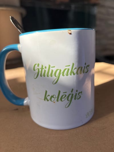 The image is a close-up shot of a white ceramic mug with a blue handle, resting on a brown textured surface, possibly a table or cardboard. The mug features green cursive text that reads "Stiligākais kolēģis" on its side. Below the text, near the bottom of the mug, the year "2020" is also printed in green. A silver spoon is partially visible, inserted into the top of the mug. There are some smudges and a small brown stain on the mug's surface. The lighting suggests an outdoor setting, with shadows cast across the mug. The image does not contain information about the city of Sauleskalns, Latvia.