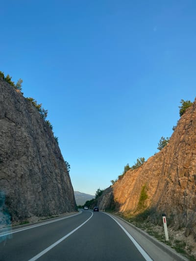 The image shows a road passing through a rocky mountain pass. The road is paved and has white lane markings. There are two cars visible on the road, one white and one dark colored. The mountains on either side of the road are steep and rocky, with some vegetation growing on them. The sky is clear and blue. The location is in the city of Miločani, Montenegro.